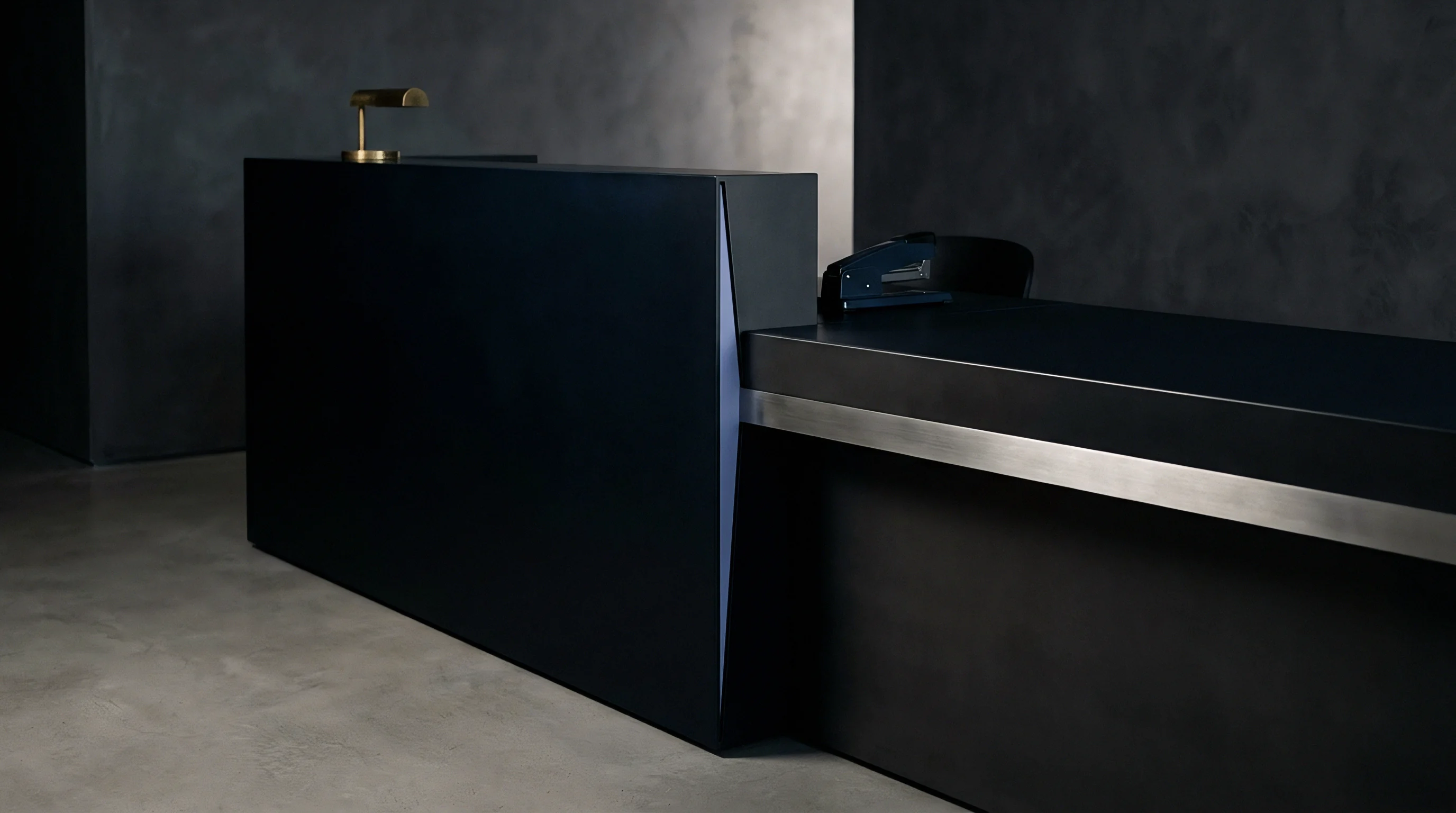 Bespoke reception desk in matte dark finish features a recessed brushed metal trim and small desk lamp positioned on a polished concrete floor.