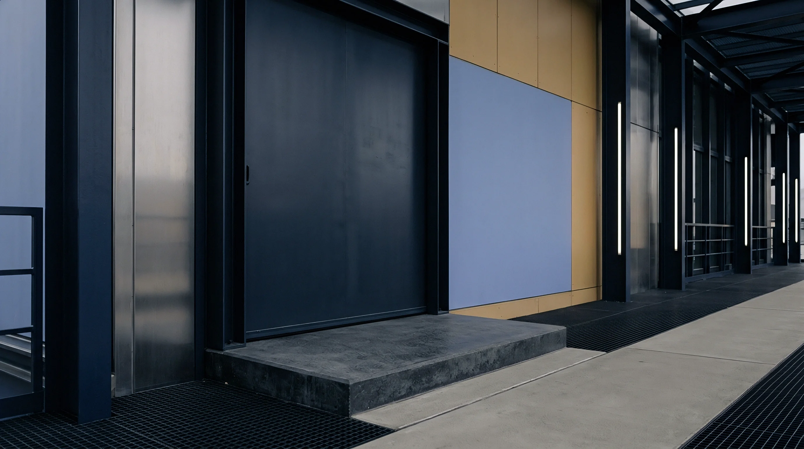 This modern industrial entryway shows a large dark metal door, concrete access steps, and metallic facade panels adjacent to a blue panel.